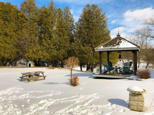 Snow-covered landscape in Schomberg featuring a rustic gazebo, Adirondack chairs, a picnic table, and tall evergreen trees.