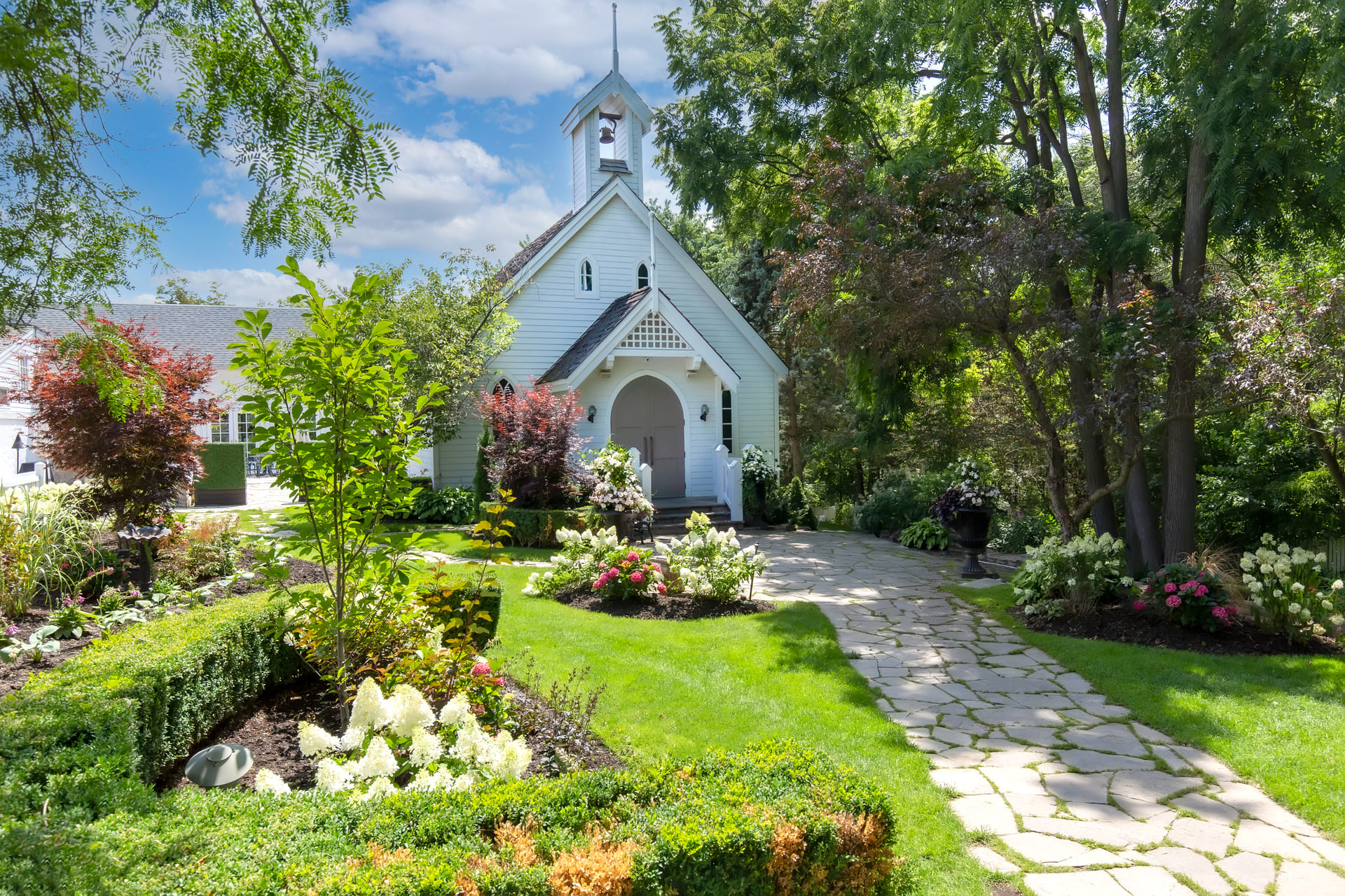 A charming white chapel with a small bell tower, surrounded by lush greenery and vibrant flower gardens, with a stone pathway leading to the entrance under a bright blue sky with scattered clouds.