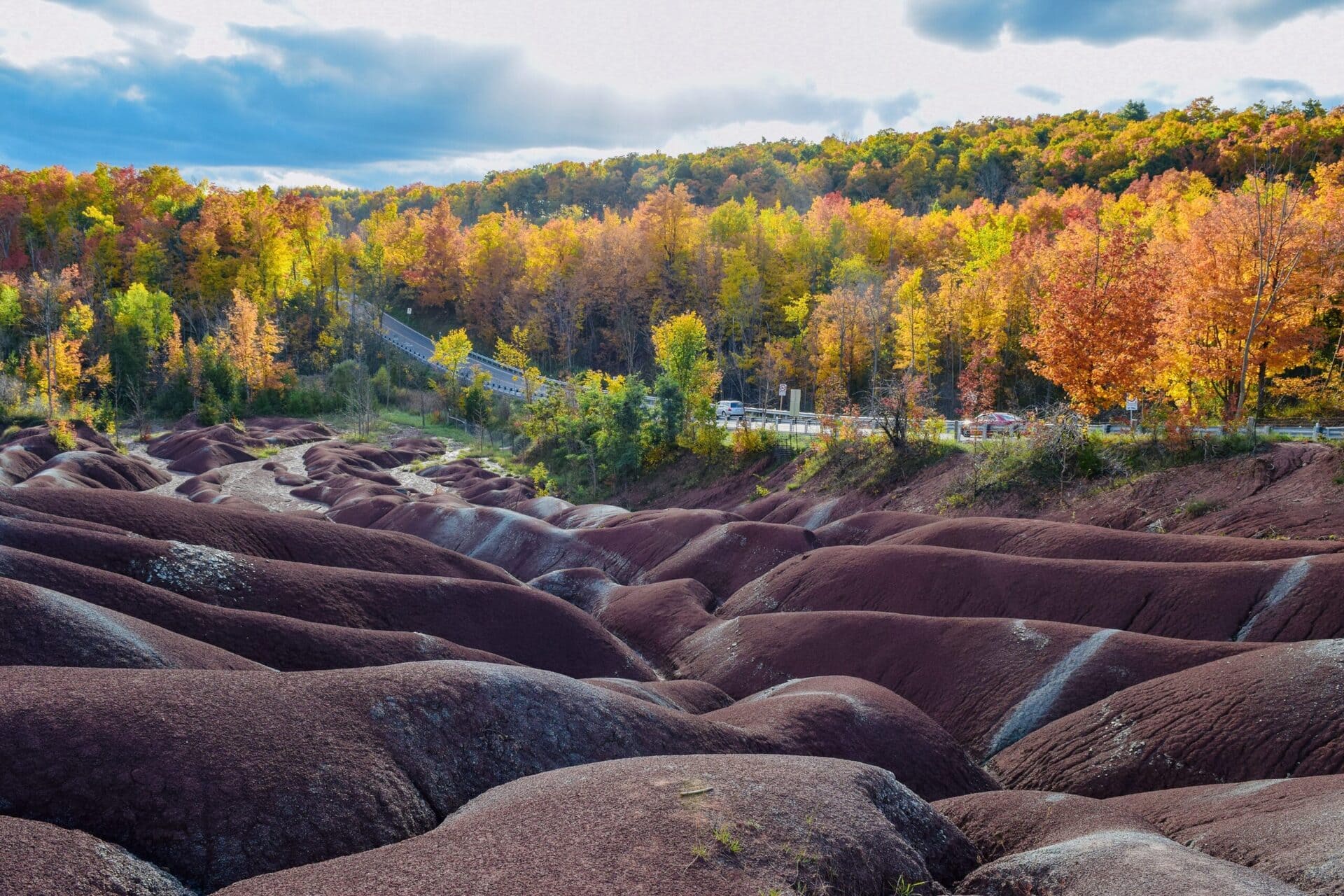 Scenic view of the Cheltenham Badlands in Caledon, Ontario, showcasing red clay hills surrounded by vibrant autumn trees and a winding road in the background.