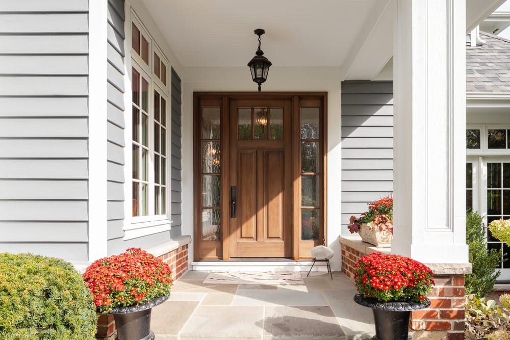 Inviting front entryway of a modern home featuring a custom wooden door with glass sidelights, light gray siding, brick accents, and seasonal planters with red flowers.