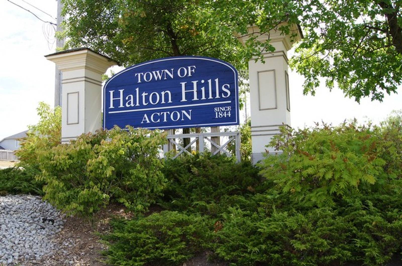 Welcome sign for the Town of Halton Hills in Acton, Ontario, featuring a blue sign with white lettering set between decorative pillars and surrounded by greenery.